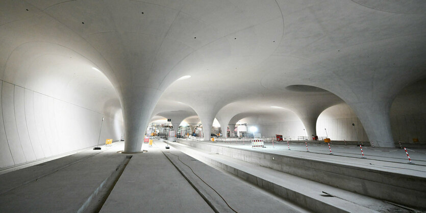 Rohbau eines unterirdischen Bahnhofs. Die Tunnelwände sind lichtgeflutet. Auf den zukünftigen Bahnsteigen steht noch Baustellenmaterial