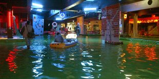 In einem Kellergeschoss steht Wasser, darauf fahren Menschen mit einem Schlauchboot. Eine Person watet durch das Wasser.