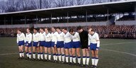 Elf männliche Fußballspieler mit weißen Trikots stehen in einem Stadion mit niedrigen Tribünen