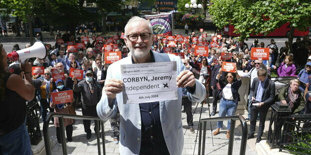 Jeremy Corbyn posiert auf einer Kundgebung: Hinter ihmstehen Menschen, die Schilder mit der Aufforderung "Vote Corbyn" hochhalten. Er trägt ein helles Jacket undl ächelt