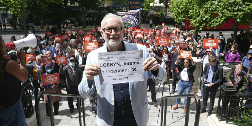 Jeremy Corbyn posiert auf einer Kundgebung: Hinter ihmstehen Menschen, die Schilder mit der Aufforderung "Vote Corbyn" hochhalten. Er trägt ein helles Jacket undl ächelt