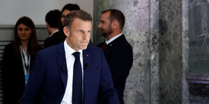 Emmanuel Macron geht mit ernstem Blick und etwas verbissenem Gesichtsausdruck an einer Gruppe von Menschen vorbei, die sich in einer Ecke unterhalten