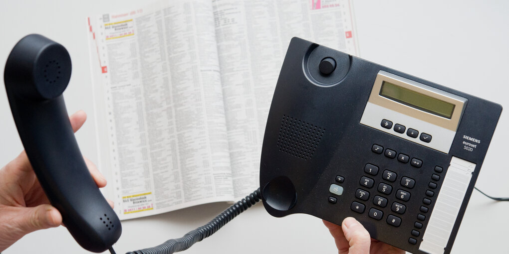 Zwei Hände halten ein Telefon und ein Telefonbuch liegt auf einem Tisch.