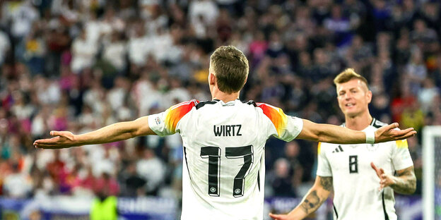 Der DfB-Spieler Florian Wirtz ist im Stadion von hinten zu sehen. Er breitet seine Arme aus. Toni Kroos kommt auf ihn zugelaufen
