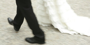 Ein Brautpaar schreitet , Mann trägt schwarzen Anzug, sie ein langes weißes Kleid, das über die Erde streift