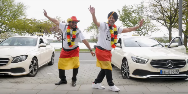 Zwei indischstämmige Männer in deutschen Nationalfarben gekleidet tanzen vor zwei Taxis
