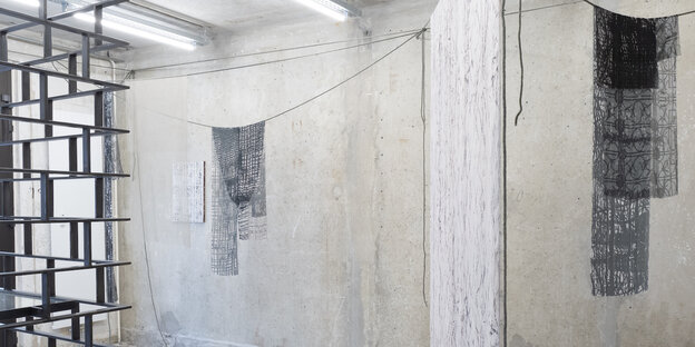 Ausstellungsansicht. Links im Bild befindet sich ein schwarzes, geschmiedetes Metallgitter. Vor den Betonwänden des Raumes sind Textilskulpturen aus durchsichtigem Stoff gespannt, auf denen leichte Muster zu erkennen sind.