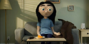 Szene aus einem animierten Film: Eine weibliche Figur aus Filz sitzt auf einem Sofa und starrt ins Leere. Sie trägt ihre schwarzen Haaren lang und hat ein hellblaues T-Shirt an.