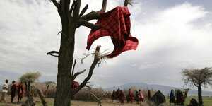Ein karriertes Tuch hängt an einem Baum und weht im Wind, im Hintergrund versammeln sich Maasai