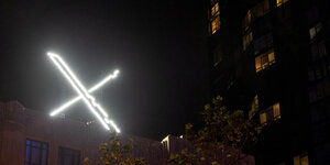leuchtendes riesiges Logo X auf einem Dach, wenige erleuchtete Fenster eines Hochhauses