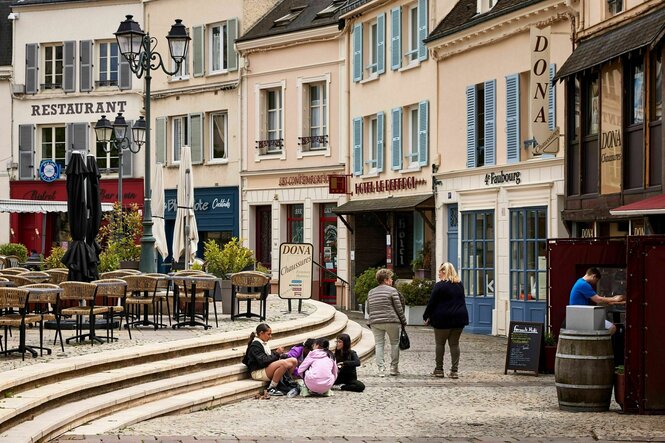 Beschauliche Stadtansicht von Dreux, kleine Geschäfte, eine Weinhandlung und die Stühle eines Cafes auf dem Marktplatz, zwei Frauen stehen bie einer Gruppe von Kindern, die auf den Stufen sitzen