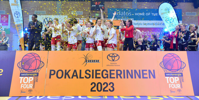 Basketballerinnen jubeln auf einer Bühne, davor steht auf einem Schild: "Pokalsiegerinnen 2023"