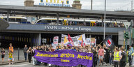 Demonstrationszug in Essen. Die Demonstrierenden tragen ein lilafarbenes Banner vor sich, auf dem in gelben Buchstaben "Zusammen gegen Rechts – Essen" steht. Im Hintergrund laufen Bahnschieben, ein Stadtmotto, "Essen – Die Folkwangstadt", ist über den Gleisen zu lesen.