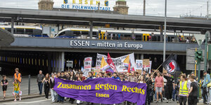 Demonstrationszug in Essen. Die Demonstrierenden tragen ein lilafarbenes Banner vor sich, auf dem in gelben Buchstaben "Zusammen gegen Rechts – Essen" steht. Im Hintergrund laufen Bahnschieben, ein Stadtmotto, "Essen – Die Folkwangstadt", ist über den Gleisen zu lesen.