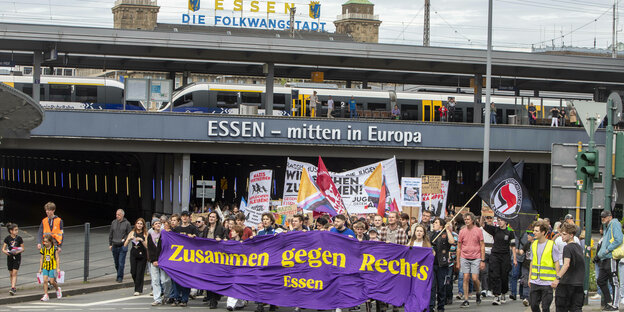 Demonstrationszug in Essen. Die Demonstrierenden tragen ein lilafarbenes Banner vor sich, auf dem in gelben Buchstaben "Zusammen gegen Rechts – Essen" steht. Im Hintergrund laufen Bahnschieben, ein Stadtmotto, "Essen – Die Folkwangstadt", ist über den Gle