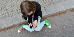 Ein Mädchen sitzt auf einem Bürgersteig und guckt auf ein Handy in seiner Hand