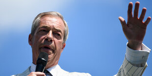 Farage mit Mikrofon hebt die Hand vor blauem Himmel