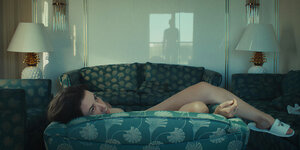 Die Protagonistin des Films liegt auf dem Sofa, in der Glasscheibe hinter ihr spiegelt sich eine männliche Figur