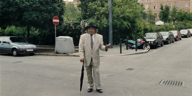 Ein Mann in einem hellen Anzug steht an einer Straßenecke in Wien auf der Straße. Er trägt einen Hut und stützt sich auf einen Regenschirm. Er spricht in die Kamera.