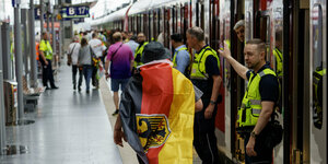 Ein Zug mit offenen Türen, an denen Ordner stehen, im Vordergrund ein in Deutschlandfahne gehüllter Fan
