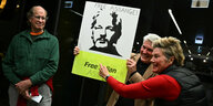 Zwei Männer und eine Frau mit einem Plakat auf das das Portrait von Julian Assange gedruckt ist