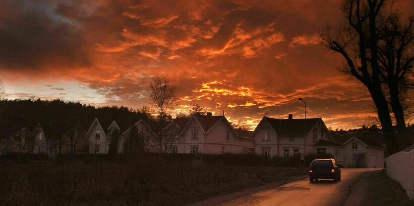 Ein Auto auf einer Landstraße fährt bei rötlich dramatischem Wolkenhimmel auf einer Häusersiedlung zu.