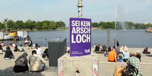 Menschen sitzen auf Stufen am Wasser, an einem Pfahl hängt ein lila Plakat mit der Aufschrift "Sei kein Arschloch".