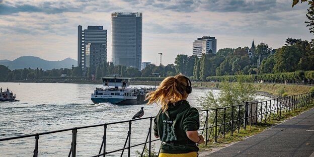 Eine Frau mit blondem Haar joggt am Rheinufer in Bonn. Ein Schiff fährt auf dem Rhein, eine Taube sitzt auf einem Geländer, die Haare der Frau wehen im Wind.