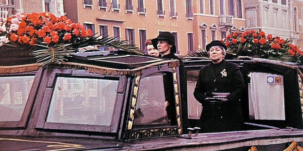 Filmstill: Zwei in Schwarz gekleidetet Frauen fahren auf einem Boot auf einem Kanal von Venedig. Das Boot ist schwarz und mit roten Rosen geschmückt.