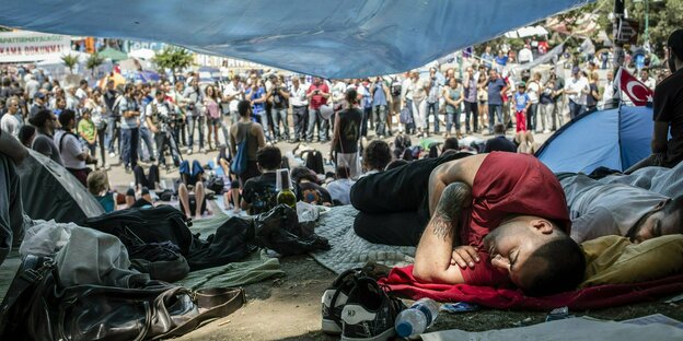 Männer liegen auf dem Boden, ein offenes Zeltdach gibt den blick frei auf eine stehende Menschenmenge