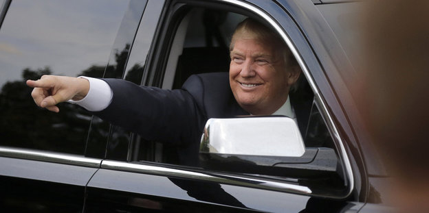 Donald Trump grinst mit ausgestrecktem Zeigefinger aus einer schwarzen Limousine.