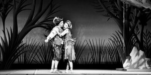 Ein Mann und eine Frau in traditioneller Kleidung mit Federn geschmückt stehen singend auf einer Bühne. Das Bühnenbild erinnert an einen dunklen Wald.