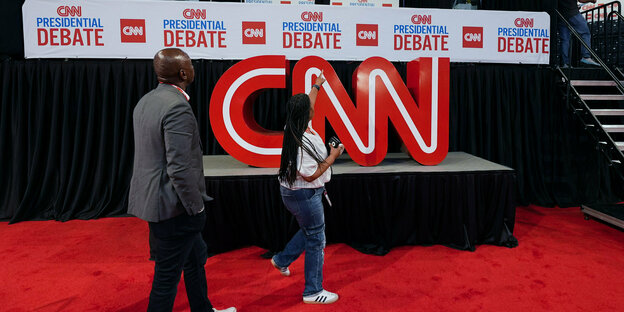 2 Mitarbeiter gehen auf rotem Teppich, oben CNN Logo, SChriftzug zur Debatte