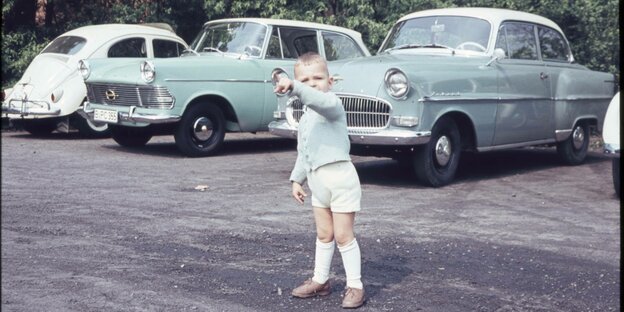 Ein Kleinkind in kurzen Hosen steht vor Autos der 50er Jahre