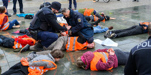 Klimaaktivisten in orangen Warnwesten liegen gefesselt auf der Straße, ein Polizist kniet auf dem Kopf und Hals einer jungen Aktivistin