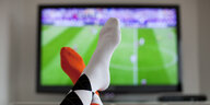 Zwei übereinander geschlagene Beine vor einem Fernseher auf dem ein Fußballspiel läuft.