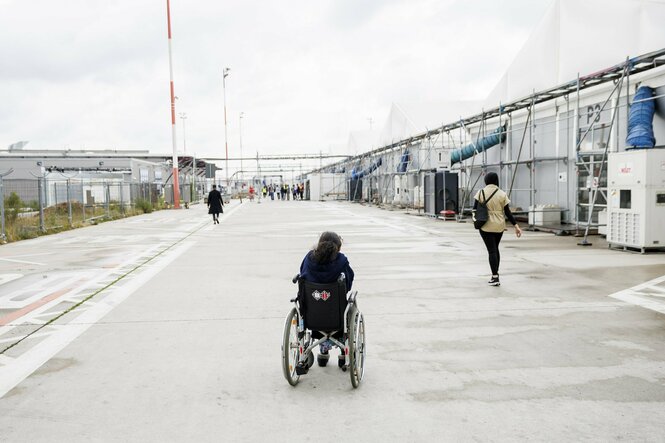 Eine Person im Rollstuhl von hinten. Der Rollstuhl steht vor dem Ankunftszentrum Tegel.