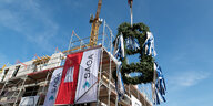 Ein Richtkranz hängt an einem Kran vor einem Rohbau, an dem das Wappen der Stadt Hamburg und ein Transparent der SAGA hängen.