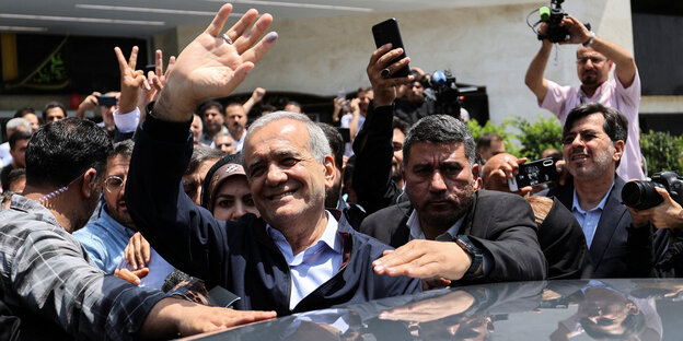 Umringt von Männern, die teils Kameras und Handys in die Höhe halten, winkt Massoud Peseschkian in die Kamera, mit der anderen Hand stützt er sich auf einem Autodach ab