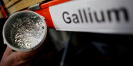 Ein Becher mit silbrig glänzendem Material neben einem Schild, auf dem steht "Gallium"