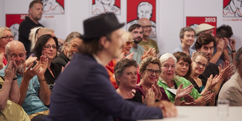 Ferdinand Schmalz sitzt im dunklen Anzug vor applaudierendem Publikum, er selbst ist auf dem Bild unscharf.