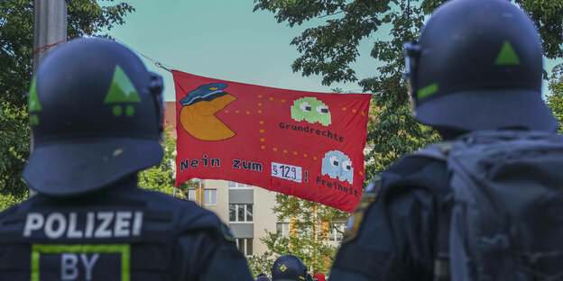 Von hinten sind zwei behelmte bayerische Polizist:innen zu sehen. Vor ihnen ist ein Transparent aufgespannt, auf dem eine Pac-Man-Figur mit Polizeimütze abgebildet ist, die kurz davor ist, die beiden Punkte "Freiheit" und "Grundrechte" zu essen. Darunter steht: „Nein zu § 129!“. Das Bild ist während der Tag-X-Proteste nach dem Urteil gegen Lina E. am 03. 06. 2023 aufgenommen worden.