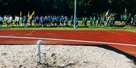 Die Mannschaften der Europeada ziehen auf das Spielfeld ein. Im Vordergrund spielt ein Kind in einer Sandgrube.