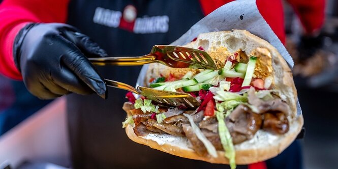 Ein Mensch hält einen Döner in der einen und eine Grillzange in der anderen Hand.