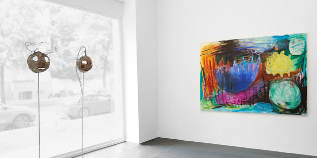 Ausstellungsräume der Galerie Tanja Wagner. An der rechten Wand hängt ein farbenreiches, abstraktes Gemälde aus Ölfarben und Ölkreiden, auf denen kräftige Grün, Blau und Gelbtöne überwiegen. Links stehen zwei runde Masken auf Metallstäben. Hinter ihnen ist das Glasfenster der Galerie zu sehen.