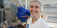 Eine Frau trägt Schutzkleidung, steht in einem Labor und lächelt