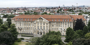 Ein großes, altes Gebäude aus der Luft fotografiert. im Vordergrund Bäume, dahinter Stadtpanorama