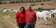 Ein kräftiger Mann legt den Arm um eine kräftige Frau, im Hintergrund eine Herde heller Kühe