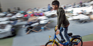 Ein Kind fährt Fahrrad in einer Turnhalle mit Schlafpritschen