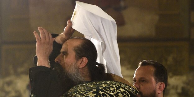 Patriarch bei der Krönung, zieht eine weiße Kopfbedeckung mit Kreuz auf